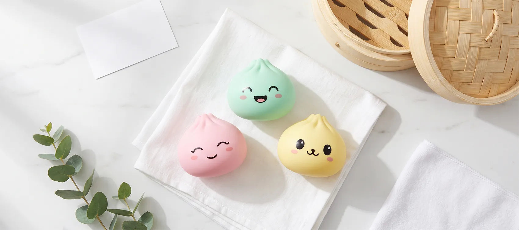 Three clean pastel dumpling squishies on a white cloth beside a bamboo steamer and eucalyptus sprig
